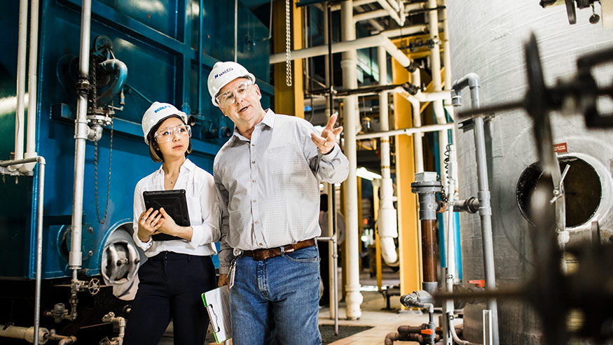 Two workers, one wearing an Ecolab hard hat and the other wearing a Nalco hard hat, speaking and gesturing while doing a walkthrough in a facility with large industrial and manufacturing equipment and carrying a tablet and checklist.