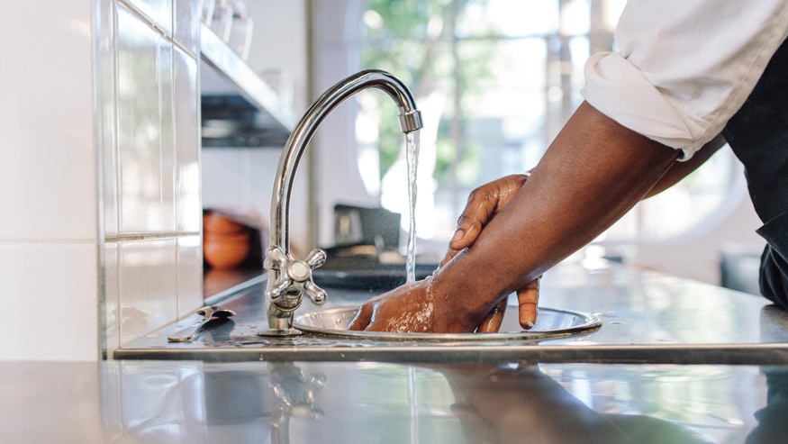 Hands being washed under a running faucet in what appears to be a restaurant or commercial kitchen.