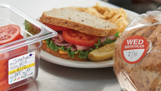 A sandwich with chips sitting on a plate with tomatoes and bread showing Daydots stickers with expiration dates.